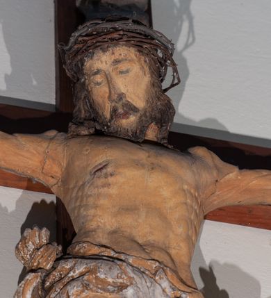 Zdjęcie nr 2: Chrystus rozpięty na krzyżu z ułożonymi niemal poziomo, szeroko rozłożonymi rękami, z nogami ugiętymi w kolanach, stopami założonymi prawa na lewą i przebitymi jednym gwoździem, z głową przechyloną na prawy bark. Twarz widoczna w trzech czwartych, owalna, z zamkniętymi oczami, okolona krótkim zarostem i spływającymi na barki włosami. Na włosy nałożona korona cierniowa. Budowa anatomiczna atletyczna, muskulatura silnie podkreślona. Karnacja ciemna, z raną w prawym boku.  Wokół bioder silnie udrapowane perizonium, przy lewym udzie z powiewającym „na wietrze” skręconym spiralnie końcem tkaniny. Nad głową Chrystusa titulus z literami IN/RI. Krzyż w kolorze naturalnego drewna, karnacja i włosy Chrystusa w kolorze naturalnym, perizonium białe i złocone, titulus biały.
