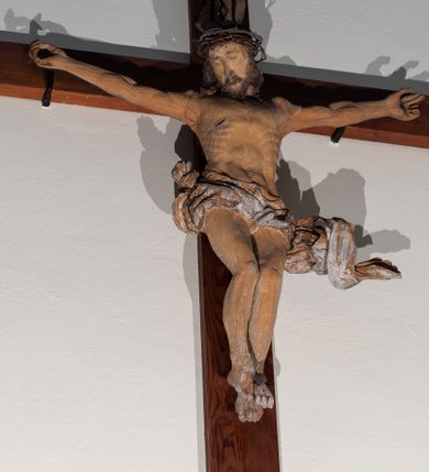 Zdjęcie nr 1: Chrystus rozpięty na krzyżu z ułożonymi niemal poziomo, szeroko rozłożonymi rękami, z nogami ugiętymi w kolanach, stopami założonymi prawa na lewą i przebitymi jednym gwoździem, z głową przechyloną na prawy bark. Twarz widoczna w trzech czwartych, owalna, z zamkniętymi oczami, okolona krótkim zarostem i spływającymi na barki włosami. Na włosy nałożona korona cierniowa. Budowa anatomiczna atletyczna, muskulatura silnie podkreślona. Karnacja ciemna, z raną w prawym boku.  Wokół bioder silnie udrapowane perizonium, przy lewym udzie z powiewającym „na wietrze” skręconym spiralnie końcem tkaniny. Nad głową Chrystusa titulus z literami IN/RI. Krzyż w kolorze naturalnego drewna, karnacja i włosy Chrystusa w kolorze naturalnym, perizonium białe i złocone, titulus biały.