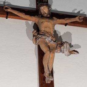 Zdjęcie nr 1: Chrystus rozpięty na krzyżu z ułożonymi niemal poziomo, szeroko rozłożonymi rękami, z nogami ugiętymi w kolanach, stopami założonymi prawa na lewą i przebitymi jednym gwoździem, z głową przechyloną na prawy bark. Twarz widoczna w trzech czwartych, owalna, z zamkniętymi oczami, okolona krótkim zarostem i spływającymi na barki włosami. Na włosy nałożona korona cierniowa. Budowa anatomiczna atletyczna, muskulatura silnie podkreślona. Karnacja ciemna, z raną w prawym boku.  Wokół bioder silnie udrapowane perizonium, przy lewym udzie z powiewającym „na wietrze” skręconym spiralnie końcem tkaniny. Nad głową Chrystusa titulus z literami IN/RI. Krzyż w kolorze naturalnego drewna, karnacja i włosy Chrystusa w kolorze naturalnym, perizonium białe i złocone, titulus biały.