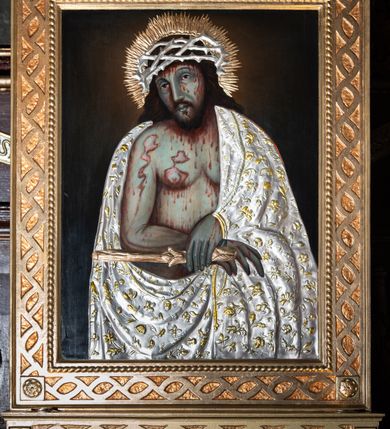 Zdjęcie nr 2: Obraz w kształcie stojącego prostokąta, w rzeźbionej złoconej ramie, na cokole, zdobionej uproszczonym motywem korony cierniowej, z rozetami w narożach. Przedstawienie w półpostaci Chrystusa w typie Ecce Homo, z głową lekko przechyloną na prawe ramię, z przegubami rąk skrępowanymi sznurem i z trzciną w lewej dłoni. Nagi korpus ze schematycznie zaznaczoną muskulaturą i obfitymi plamami krwi okryty metalową srebrzystą sukienką ze złoconymi motywami roślinnymi. Na włosach metalowa ażurowa korona cierniowa upleciona z cienkich gałązek ciernia. Wokół głowy złocisty nimb promienisty. Tło ciemne monochromatyczne. 