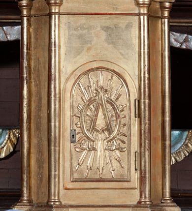 Zdjęcie nr 1: Prostopadłościenna szafka na rzucie prostokąta o ściętych od przodu narożnikach i od przodu opięta wysmukłymi kolumienkami o gładkich trzonach i kielichowych kapitelach, ustawionych na cokole i podtrzymujących profilowany gzyms w zwieńczeniu. W przedniej ściance prostokątne zamknięte półkoliście drzwiczki, osadzone na zawiasach i zamykane na zamek, wypełnione płaskorzeźbionymi literami A i Ω na tle promieni. Całe tabernakulum pozłocone.