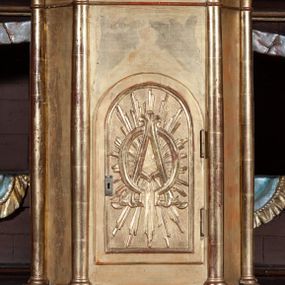 Zdjęcie nr 1: Prostopadłościenna szafka na rzucie prostokąta o ściętych od przodu narożnikach i od przodu opięta wysmukłymi kolumienkami o gładkich trzonach i kielichowych kapitelach, ustawionych na cokole i podtrzymujących profilowany gzyms w zwieńczeniu. W przedniej ściance prostokątne zamknięte półkoliście drzwiczki, osadzone na zawiasach i zamykane na zamek, wypełnione płaskorzeźbionymi literami A i Ω na tle promieni. Całe tabernakulum pozłocone.