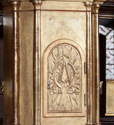 Zdjęcie nr 2: Prostopadłościenna szafka na rzucie prostokąta o ściętych od przodu narożnikach i od przodu opięta wysmukłymi kolumienkami o gładkich trzonach i kielichowych kapitelach, ustawionych na cokole i podtrzymujących profilowany gzyms w zwieńczeniu. W przedniej ściance prostokątne zamknięte półkoliście drzwiczki, osadzone na zawiasach i zamykane na zamek, wypełnione płaskorzeźbionymi literami A i Ω na tle promieni. Całe tabernakulum pozłocone.