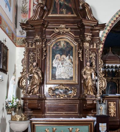 Zdjęcie nr 2: Ołtarz architektoniczny, jednokondygnacyjny, jednoosiowy ze zwieńczeniem. Antepedium z płaskorzeźbionym krzyżem ujętym wolutami z motywami lilii. Nastawę ujmują ukośnie ustawione zwielokrotnione  pilastry z konsolami, na których stoją figury. Pilastry podtrzymują belkowanie, które nad zewnętrznymi pilastrami jest lekko wklęsłe, nad wewnętrznymi zwieńczone falującymi naczółkami, a nad osią środkową  z lekko wybrzuszonym architrawem. Zwieńczenie o lekko wklęsłych bokach zamknięte profilowanym gzymsem w formie łuku nadwieszonego. W polu środkowym wnęka w kształcie stojącego prostokąta zakończonego łukiem odcinkowym, nadwieszonym, w niej obraz &quot;Św. Anna Samotrzeć&quot; i &quot;Męczeństwo św. Sebastiana&quot; na zasuwie. Pole obrazowe w zwieńczeniu powtarza jego kształt, wypełnione wizerunkiem św. Floriana. W partii predelli wkomponowano fantazyjny kartusz z obrazem przedstawiającym hołd Trzech Króli. Na dekorację ornamentalną składają się motywy rocaille&#039;owe na pilastrach, uszy ujmujące nastawę z rocaille&#039;ami w części wypełnionymi kratką regencyjną, kartusz w partii belkowania oraz drobne motywy roślinne i rokokowe w partii zwieńczenia., 

