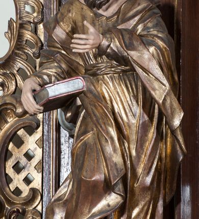 Zdjęcie nr 3: Rzeźba pełnoplastyczna, przyścienna, przedstawiająca w całej postaci nierozpoznanego proroka (Izajasza?). Prorok stoi na niskim prostopadłościennym cokole, w kontrapoście, w prawą nogą ugiętą w kolanie i bosą stopą wysuniętą do przodu. Sylwetka esowato wygięta, z głową lekko pochyloną i zwróconą w lewo. Twarz pociągła, szczupła, okolona ciemnymi włosami i zarostem. Ręce ugięte w łokciach, prawą podtrzymuje zamkniętą księgę opartą o biodro. Lewą dłoń wyciąga do przodu w sugestywnym geście. Ubrany w długą złotą tunikę i płaszcz, przewiązane w pasie o fałdach kształtowanych kanciasto i dynamicznie.