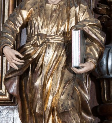 Zdjęcie nr 6: Rzeźba pełnoplastyczna, przyścienna, przedstawiająca w całej postaci nierozpoznanego proroka (Izajasza?). Prorok stoi na niskim prostopadłościennym cokole, w kontrapoście, w prawą nogą ugiętą w kolanie i bosą stopą wysuniętą do przodu. Sylwetka esowato wygięta, z głową lekko pochyloną i zwróconą w lewo. Twarz pociągła, szczupła, okolona ciemnymi włosami i zarostem. Ręce ugięte w łokciach, prawą podtrzymuje zamkniętą księgę opartą o biodro. Lewą dłoń wyciąga do przodu w sugestywnym geście. Ubrany w długą złotą tunikę i płaszcz, przewiązane w pasie o fałdach kształtowanych kanciasto i dynamicznie.