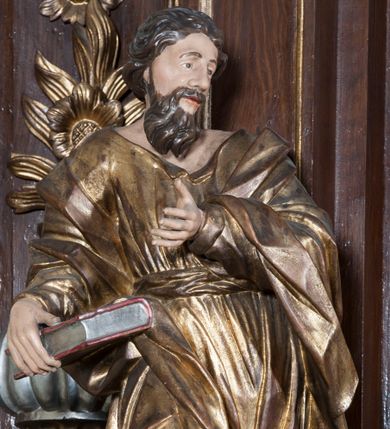 Zdjęcie nr 1: Rzeźba pełnoplastyczna, przyścienna, przedstawiająca w całej postaci nierozpoznanego proroka (Izajasza?). Prorok stoi na niskim prostopadłościennym cokole, w kontrapoście, w prawą nogą ugiętą w kolanie i bosą stopą wysuniętą do przodu. Sylwetka esowato wygięta, z głową lekko pochyloną i zwróconą w lewo. Twarz pociągła, szczupła, okolona ciemnymi włosami i zarostem. Ręce ugięte w łokciach, prawą podtrzymuje zamkniętą księgę opartą o biodro. Lewą dłoń wyciąga do przodu w sugestywnym geście. Ubrany w długą złotą tunikę i płaszcz, przewiązane w pasie o fałdach kształtowanych kanciasto i dynamicznie.