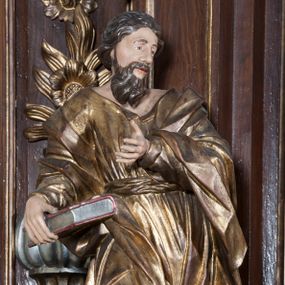 Zdjęcie nr 1: Rzeźba pełnoplastyczna, przyścienna, przedstawiająca w całej postaci nierozpoznanego proroka (Izajasza?). Prorok stoi na niskim prostopadłościennym cokole, w kontrapoście, w prawą nogą ugiętą w kolanie i bosą stopą wysuniętą do przodu. Sylwetka esowato wygięta, z głową lekko pochyloną i zwróconą w lewo. Twarz pociągła, szczupła, okolona ciemnymi włosami i zarostem. Ręce ugięte w łokciach, prawą podtrzymuje zamkniętą księgę opartą o biodro. Lewą dłoń wyciąga do przodu w sugestywnym geście. Ubrany w długą złotą tunikę i płaszcz, przewiązane w pasie o fałdach kształtowanych kanciasto i dynamicznie.