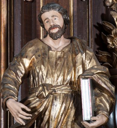 Zdjęcie nr 8: Rzeźba pełnoplastyczna, przyścienna, przedstawiająca w całej postaci nierozpoznanego proroka (Izajasza?). Prorok stoi na niskim prostopadłościennym cokole, w kontrapoście, w prawą nogą ugiętą w kolanie i bosą stopą wysuniętą do przodu. Sylwetka esowato wygięta, z głową lekko pochyloną i zwróconą w lewo. Twarz pociągła, szczupła, okolona ciemnymi włosami i zarostem. Ręce ugięte w łokciach, prawą podtrzymuje zamkniętą księgę opartą o biodro. Lewą dłoń wyciąga do przodu w sugestywnym geście. Ubrany w długą złotą tunikę i płaszcz, przewiązane w pasie o fałdach kształtowanych kanciasto i dynamicznie.