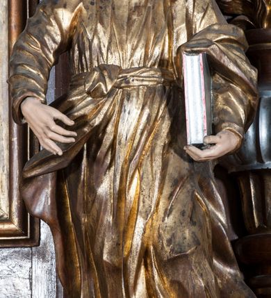 Zdjęcie nr 5: Rzeźba pełnoplastyczna, przyścienna, przedstawiająca w całej postaci nierozpoznanego proroka (Izajasza?). Prorok stoi na niskim prostopadłościennym cokole, w kontrapoście, w prawą nogą ugiętą w kolanie i bosą stopą wysuniętą do przodu. Sylwetka esowato wygięta, z głową lekko pochyloną i zwróconą w lewo. Twarz pociągła, szczupła, okolona ciemnymi włosami i zarostem. Ręce ugięte w łokciach, prawą podtrzymuje zamkniętą księgę opartą o biodro. Lewą dłoń wyciąga do przodu w sugestywnym geście. Ubrany w długą złotą tunikę i płaszcz, przewiązane w pasie o fałdach kształtowanych kanciasto i dynamicznie.