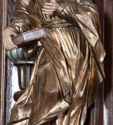 Zdjęcie nr 2: Rzeźba pełnoplastyczna, przyścienna, przedstawiająca w całej postaci nierozpoznanego proroka (Izajasza?). Prorok stoi na niskim prostopadłościennym cokole, w kontrapoście, w prawą nogą ugiętą w kolanie i bosą stopą wysuniętą do przodu. Sylwetka esowato wygięta, z głową lekko pochyloną i zwróconą w lewo. Twarz pociągła, szczupła, okolona ciemnymi włosami i zarostem. Ręce ugięte w łokciach, prawą podtrzymuje zamkniętą księgę opartą o biodro. Lewą dłoń wyciąga do przodu w sugestywnym geście. Ubrany w długą złotą tunikę i płaszcz, przewiązane w pasie o fałdach kształtowanych kanciasto i dynamicznie.