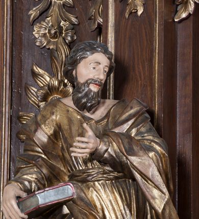 Zdjęcie nr 7: Rzeźba pełnoplastyczna, przyścienna, przedstawiająca w całej postaci nierozpoznanego proroka (Izajasza?). Prorok stoi na niskim prostopadłościennym cokole, w kontrapoście, w prawą nogą ugiętą w kolanie i bosą stopą wysuniętą do przodu. Sylwetka esowato wygięta, z głową lekko pochyloną i zwróconą w lewo. Twarz pociągła, szczupła, okolona ciemnymi włosami i zarostem. Ręce ugięte w łokciach, prawą podtrzymuje zamkniętą księgę opartą o biodro. Lewą dłoń wyciąga do przodu w sugestywnym geście. Ubrany w długą złotą tunikę i płaszcz, przewiązane w pasie o fałdach kształtowanych kanciasto i dynamicznie.