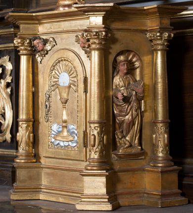 Zdjęcie nr 1: Prostopadłościenna szafka na rzucie prostokąta o ściętych od przodu narożnikach, na cokole, od przodu opięta kolumienkami o trzonach w jednej trzeciej wysokości zdobionej ornamentem okuciowym, z kompozytowymi kapitelami, ustawionymi na cokołach i wysuniętych postumentach. Kolumny podtrzymują uproszczone belkowanie i profilowany gzyms występujące nad nimi. W przedniej ściance prostokątne zamknięte półkoliście drzwiczki, osadzone na zawiasach i zamykane na zamek, ozdobione płyciną powtarzającą kształt drzwiczek, z płaskorzeźbionym ustawionym na korporale kielichem mszalnym z hostią na tle promieni. W górnych narożach przedniej ściany uskrzydlone główki aniołków. W polach pomiędzy kolumnami płaskorzeźbione postacie nieokreślonych świętych. Tabernakulum pozłocone, hostia i korporał posrebrzone.