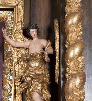 Zdjęcie nr 2: Rzeźby pełnoplastyczne, przyścienne wyobrażające dwóch aniołów ze skrzydłami, ukazanych w całej postaci, w antytetycznych pozach, w kontrapoście, w trzech czwartych zwróconych ku środkowi ołtarza, z głowami skierowanymi do widza. Anioł po prawej prawą ręką uniesioną na wysokość barku, a anioł po lewej lewą ręką wskazują wymownym gestem na obraz Matki Boskiej umieszczony w retabulum. Obaj aniołowie o młodzieńczych twarzach okolonych ciemnymi kędzierzawymi włosami. Ramiona i tors nagie, o jasnej karnacji. Brzuch, biodra i uda okryte silnie pofałdowaną dynamicznie złotą draperią spływającą z tyłu postaci aż do ziemi. Na nogach wysokie buty odsłaniające palce.  Pod stopami srebrzyste laserowane na niebiesko obłoki.