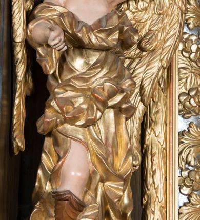 Zdjęcie nr 6: Rzeźby pełnoplastyczne, przyścienne wyobrażające dwóch aniołów ze skrzydłami, ukazanych w całej postaci, w antytetycznych pozach, w kontrapoście, w trzech czwartych zwróconych ku środkowi ołtarza, z głowami skierowanymi do widza. Anioł po prawej prawą ręką uniesioną na wysokość barku, a anioł po lewej lewą ręką wskazują wymownym gestem na obraz Matki Boskiej umieszczony w retabulum. Obaj aniołowie o młodzieńczych twarzach okolonych ciemnymi kędzierzawymi włosami. Ramiona i tors nagie, o jasnej karnacji. Brzuch, biodra i uda okryte silnie pofałdowaną dynamicznie złotą draperią spływającą z tyłu postaci aż do ziemi. Na nogach wysokie buty odsłaniające palce.  Pod stopami srebrzyste laserowane na niebiesko obłoki.