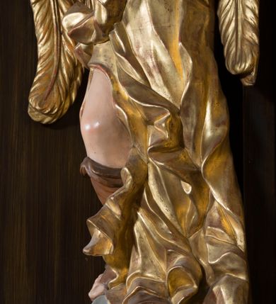 Zdjęcie nr 9: Rzeźby pełnoplastyczne, przyścienne wyobrażające dwóch aniołów ze skrzydłami, ukazanych w całej postaci, w antytetycznych pozach, w kontrapoście, w trzech czwartych zwróconych ku środkowi ołtarza, z głowami skierowanymi do widza. Anioł po prawej prawą ręką uniesioną na wysokość barku, a anioł po lewej lewą ręką wskazują wymownym gestem na obraz Matki Boskiej umieszczony w retabulum. Obaj aniołowie o młodzieńczych twarzach okolonych ciemnymi kędzierzawymi włosami. Ramiona i tors nagie, o jasnej karnacji. Brzuch, biodra i uda okryte silnie pofałdowaną dynamicznie złotą draperią spływającą z tyłu postaci aż do ziemi. Na nogach wysokie buty odsłaniające palce.  Pod stopami srebrzyste laserowane na niebiesko obłoki.