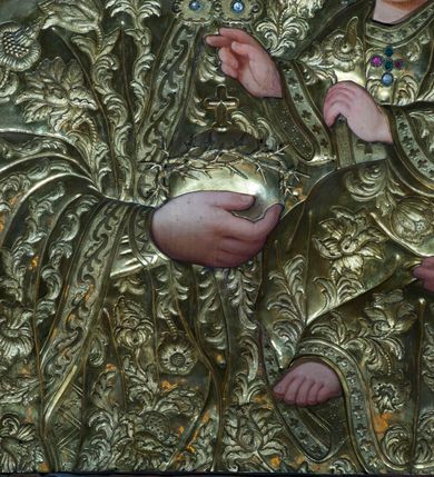 Zdjęcie nr 4: Obraz w kształcie stojącego prostokąta, w rzeźbionej, złoconej i srebrzonej ramie z motywem liści. Przedstawienie Matki Boskiej z Dzieciątkiem, zwanej Matką Boską Lanckorońską, w typie Hodegetrii. Maria ukazana w półpostaci , frontalnie, z rękami zgiętymi w łokciach, lewą podtrzymuje siedzącego na jej przedramieniu Jezusa, prawa dłoń złożona na piersi. Głowa Marii nieduża, osadzona na smukłej szyi; twarz o jasnej karnacji, delikatnych rysach, dużych niebieskich oczach zwróconych na wprost oraz małych ustach. Jezus, ukazany bokiem, zwrócony w lewo, z twarzą widoczną w trzech czwartych, prawą ręką błogosławi, w lewej dłoni trzyma niewielką książeczkę. Maria i Jezus okryci metalowymi srebrzonymi (?) sukienkami zdobionymi motywami roślinnymi; na głowach metalowe korony zamknięte, wokół głów nimby promieniste oraz dwanaście gwiazd. Tło ciemne, monochromatyczne.
