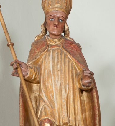 Zdjęcie nr 1: Rzeźba pełnoplastyczna, przyścienna, ścięta z tyłu. Święty ukazany w całej postaci, frontalnie, stoi w kontrapoście. Ręce ugięte w łokciach. W lewej dłoni trzyma ferulę, prawą trzyma drzewce dużego krzyża. Twarz kwadratowa, o wyraźnej linii żuchwy, małych oczach, prostym nosie i niewielkich ustach. Święty ubrany w albę, komżę, spiętą pod szyją kapę, na głowie infuła. Polichromia naturalistyczna w partiach ciała, szaty złocone.
