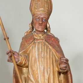 Zdjęcie nr 1: Rzeźba pełnoplastyczna, przyścienna, ścięta z tyłu. Święty ukazany w całej postaci, frontalnie, stoi w kontrapoście. Ręce ugięte w łokciach. W lewej dłoni trzyma ferulę, prawą trzyma drzewce dużego krzyża. Twarz kwadratowa, o wyraźnej linii żuchwy, małych oczach, prostym nosie i niewielkich ustach. Święty ubrany w albę, komżę, spiętą pod szyją kapę, na głowie infuła. Polichromia naturalistyczna w partiach ciała, szaty złocone.