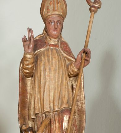 Zdjęcie nr 3: Rzeźba pełnoplastyczna, przyścienna, ścięta z tyłu. Święty, ukazany frontalnie, stoi z rękami ugiętymi w łokciach, prawą błogosławi, lewą podtrzymuje pastorał. Twarz kwadratowa, o wyraźnej linii żuchwy, małych oczach, prostym nosie i niewielkich ustach. Święty ubrany w długą albę, komżę, spiętą pod szyją kapę, na głowie infułą ozdobiona dużym krzyżem. Polichromia naturalistyczna w partiach ciała, szaty złocone.