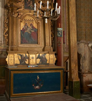 Zdjęcie nr 2: Obraz w formie stojącego prostokąta z przedstawieniem św. Józefa z Dzieciątkiem. Święty ukazany w półpostaci, frontalnie, w pozycji siedzącej, z głową zwróconą w lewo. Prawą dłonią trzyma rączkę Dzieciątka z gałązką lilii, lewą obejmuje je. Twarz ma pociągłą, o małych, migdałowatych oczach, niewielkim nosie i pełnych ustach, okoloną gęstą, krótką brodą oraz krótkimi, brązowymi włosami. Święty ubrany jest w fioletową szatę zapinaną na guziki oraz brązowy płaszcz z zieloną podszewką zarzucony na kolana i lewe ramię. Dzieciątko ukazane w pozycji stojącej, zwrócone w trzech czwartych w prawo, z głową skierowaną frontalnie, opiera się o ramię Józefa, lewą rękę wyciąga w bok, w dłoni trzyma lilię. Twarz ma okrągłą, o migdałowatych oczach, małym nosie i pełnych ustach, okoloną jasnymi, krótkimi, kręconymi włosami. W partii bioder przesłonięte białą tkaniną. Tło jednolite, ciemnobrązowe. Obraz ujęty jest profilowaną, złoconą ramą.