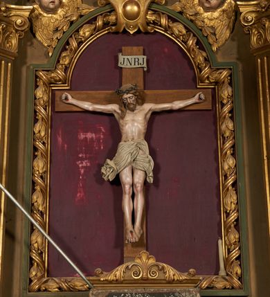 Zdjęcie nr 3: Nastawa ołtarzowa architektoniczna, jednoosiowa, jednokondygnacyjna ze zwieńczeniem. Ołtarz drewniany, prostopadłościenny, mensa drewniana. Nastawa ustawiona na cokole zakończonym gzymsem z motywem wolego oka, dekorowanym płycinami z kartuszami, z wyodrębnionym po środku tabernakulum flankowanym pilastrami z konsolami i kampanulami. Drzwiczki tabernakulum w formie stojącego prostokąta zamkniętego łukiem półkolistym z przedstawieniem kielicha z hostią w glorii. Nastawa ujęta parą zdwojonych filarów ustawionych pod kątem, o kanelowanych trzonach i kapitelach kompozytowych dźwigających fragmenty przerwanego belkowania dekorowanego wolim okiem oraz ćwierćkolistego, przerwanego przyczółka, na których siedzi para puttów. Na tle filarów, po zewnętrznej stronie ustawione są figury biskupów. W polu środkowym wnęka w formie stojącego prostokąta zamkniętego łukiem półkolistym nadwieszonym, ujęta grubą ramą dekorowaną wstęgą przeplataną z pąkami kwiatów, zwieńczona kartuszem; w polu figura Chrystusa Ukrzyżowanego na czerwonym tle. W przyłuczach dwie uskrzydlone główki anielskie. Zwieńczenie w formie stojącego prostokąta ujętego spływami wolutowymi, zamknięte fragmentem gzymsu, na którym znajduje się szczyt utworzony z przeciwstawnie zwróconych wolut z anthemionem. W polu zwieńczenia obraz w formie stojącego prostokąta z zaokrąglonymi górnymi narożami z przedstawieniem Matki Boskiej Bolesnej, ujęty profilowaną ramą. Ponad nim wić roślinna. Struktura ołtarza malowana na kolor jasnobrązowy, profile i ornamenty złocone.
