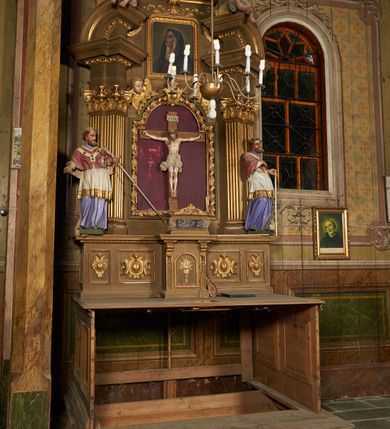 Zdjęcie nr 2: Nastawa ołtarzowa architektoniczna, jednoosiowa, jednokondygnacyjna ze zwieńczeniem. Ołtarz drewniany, prostopadłościenny, mensa drewniana. Nastawa ustawiona na cokole zakończonym gzymsem z motywem wolego oka, dekorowanym płycinami z kartuszami, z wyodrębnionym po środku tabernakulum flankowanym pilastrami z konsolami i kampanulami. Drzwiczki tabernakulum w formie stojącego prostokąta zamkniętego łukiem półkolistym z przedstawieniem kielicha z hostią w glorii. Nastawa ujęta parą zdwojonych filarów ustawionych pod kątem, o kanelowanych trzonach i kapitelach kompozytowych dźwigających fragmenty przerwanego belkowania dekorowanego wolim okiem oraz ćwierćkolistego, przerwanego przyczółka, na których siedzi para puttów. Na tle filarów, po zewnętrznej stronie ustawione są figury biskupów. W polu środkowym wnęka w formie stojącego prostokąta zamkniętego łukiem półkolistym nadwieszonym, ujęta grubą ramą dekorowaną wstęgą przeplataną z pąkami kwiatów, zwieńczona kartuszem; w polu figura Chrystusa Ukrzyżowanego na czerwonym tle. W przyłuczach dwie uskrzydlone główki anielskie. Zwieńczenie w formie stojącego prostokąta ujętego spływami wolutowymi, zamknięte fragmentem gzymsu, na którym znajduje się szczyt utworzony z przeciwstawnie zwróconych wolut z anthemionem. W polu zwieńczenia obraz w formie stojącego prostokąta z zaokrąglonymi górnymi narożami z przedstawieniem Matki Boskiej Bolesnej, ujęty profilowaną ramą. Ponad nim wić roślinna. Struktura ołtarza malowana na kolor jasnobrązowy, profile i ornamenty złocone.
