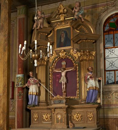 Zdjęcie nr 1: Nastawa ołtarzowa architektoniczna, jednoosiowa, jednokondygnacyjna ze zwieńczeniem. Ołtarz drewniany, prostopadłościenny, mensa drewniana. Nastawa ustawiona na cokole zakończonym gzymsem z motywem wolego oka, dekorowanym płycinami z kartuszami, z wyodrębnionym po środku tabernakulum flankowanym pilastrami z konsolami i kampanulami. Drzwiczki tabernakulum w formie stojącego prostokąta zamkniętego łukiem półkolistym z przedstawieniem kielicha z hostią w glorii. Nastawa ujęta parą zdwojonych filarów ustawionych pod kątem, o kanelowanych trzonach i kapitelach kompozytowych dźwigających fragmenty przerwanego belkowania dekorowanego wolim okiem oraz ćwierćkolistego, przerwanego przyczółka, na których siedzi para puttów. Na tle filarów, po zewnętrznej stronie ustawione są figury biskupów. W polu środkowym wnęka w formie stojącego prostokąta zamkniętego łukiem półkolistym nadwieszonym, ujęta grubą ramą dekorowaną wstęgą przeplataną z pąkami kwiatów, zwieńczona kartuszem; w polu figura Chrystusa Ukrzyżowanego na czerwonym tle. W przyłuczach dwie uskrzydlone główki anielskie. Zwieńczenie w formie stojącego prostokąta ujętego spływami wolutowymi, zamknięte fragmentem gzymsu, na którym znajduje się szczyt utworzony z przeciwstawnie zwróconych wolut z anthemionem. W polu zwieńczenia obraz w formie stojącego prostokąta z zaokrąglonymi górnymi narożami z przedstawieniem Matki Boskiej Bolesnej, ujęty profilowaną ramą. Ponad nim wić roślinna. Struktura ołtarza malowana na kolor jasnobrązowy, profile i ornamenty złocone.
