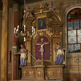 Zdjęcie nr 1: Nastawa ołtarzowa architektoniczna, jednoosiowa, jednokondygnacyjna ze zwieńczeniem. Ołtarz drewniany, prostopadłościenny, mensa drewniana. Nastawa ustawiona na cokole zakończonym gzymsem z motywem wolego oka, dekorowanym płycinami z kartuszami, z wyodrębnionym po środku tabernakulum flankowanym pilastrami z konsolami i kampanulami. Drzwiczki tabernakulum w formie stojącego prostokąta zamkniętego łukiem półkolistym z przedstawieniem kielicha z hostią w glorii. Nastawa ujęta parą zdwojonych filarów ustawionych pod kątem, o kanelowanych trzonach i kapitelach kompozytowych dźwigających fragmenty przerwanego belkowania dekorowanego wolim okiem oraz ćwierćkolistego, przerwanego przyczółka, na których siedzi para puttów. Na tle filarów, po zewnętrznej stronie ustawione są figury biskupów. W polu środkowym wnęka w formie stojącego prostokąta zamkniętego łukiem półkolistym nadwieszonym, ujęta grubą ramą dekorowaną wstęgą przeplataną z pąkami kwiatów, zwieńczona kartuszem; w polu figura Chrystusa Ukrzyżowanego na czerwonym tle. W przyłuczach dwie uskrzydlone główki anielskie. Zwieńczenie w formie stojącego prostokąta ujętego spływami wolutowymi, zamknięte fragmentem gzymsu, na którym znajduje się szczyt utworzony z przeciwstawnie zwróconych wolut z anthemionem. W polu zwieńczenia obraz w formie stojącego prostokąta z zaokrąglonymi górnymi narożami z przedstawieniem Matki Boskiej Bolesnej, ujęty profilowaną ramą. Ponad nim wić roślinna. Struktura ołtarza malowana na kolor jasnobrązowy, profile i ornamenty złocone.
