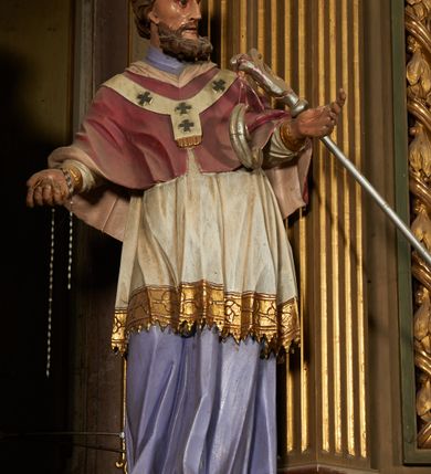 Zdjęcie nr 4: Nastawa ołtarzowa architektoniczna, jednoosiowa, jednokondygnacyjna ze zwieńczeniem. Ołtarz drewniany, prostopadłościenny, mensa drewniana. Nastawa ustawiona na cokole zakończonym gzymsem z motywem wolego oka, dekorowanym płycinami z kartuszami, z wyodrębnionym po środku tabernakulum flankowanym pilastrami z konsolami i kampanulami. Drzwiczki tabernakulum w formie stojącego prostokąta zamkniętego łukiem półkolistym z przedstawieniem kielicha z hostią w glorii. Nastawa ujęta parą zdwojonych filarów ustawionych pod kątem, o kanelowanych trzonach i kapitelach kompozytowych dźwigających fragmenty przerwanego belkowania dekorowanego wolim okiem oraz ćwierćkolistego, przerwanego przyczółka, na których siedzi para puttów. Na tle filarów, po zewnętrznej stronie ustawione są figury biskupów. W polu środkowym wnęka w formie stojącego prostokąta zamkniętego łukiem półkolistym nadwieszonym, ujęta grubą ramą dekorowaną wstęgą przeplataną z pąkami kwiatów, zwieńczona kartuszem; w polu figura Chrystusa Ukrzyżowanego na czerwonym tle. W przyłuczach dwie uskrzydlone główki anielskie. Zwieńczenie w formie stojącego prostokąta ujętego spływami wolutowymi, zamknięte fragmentem gzymsu, na którym znajduje się szczyt utworzony z przeciwstawnie zwróconych wolut z anthemionem. W polu zwieńczenia obraz w formie stojącego prostokąta z zaokrąglonymi górnymi narożami z przedstawieniem Matki Boskiej Bolesnej, ujęty profilowaną ramą. Ponad nim wić roślinna. Struktura ołtarza malowana na kolor jasnobrązowy, profile i ornamenty złocone.
