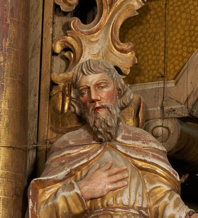 Zdjęcie nr 4: Figura drążona z tyłu, ustawiona na podstawie na nieregularnym planie. Święty ukazany frontalnie, z pochyloną głową skierowaną w prawo, prawa noga oparta na postumencie. Lewa ręka ugięta, wyciągnięta w bok, prawa złożona na piersi. Twarz ma podłużną, o wąskich, migdałowatych oczach, dużym nosie i wąskich, rozchylonych ustach, okoloną falowaną brodą oraz krótkimi włosami z grzywką nad czołem. Święty ubrany w złoconą, przylegającą do ciała, fałdowaną tunikę, przepasaną w talii oraz złocony płaszcz zapięty na piersi, opadający na ramiona z zarzuconą połą na prawą nogę. Polichromia ciała naturalistyczna.

