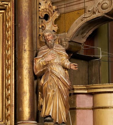 Zdjęcie nr 2: Figura drążona z tyłu, ustawiona na podstawie na nieregularnym planie. Święty ukazany frontalnie, z pochyloną głową skierowaną w prawo, prawa noga oparta na postumencie. Lewa ręka ugięta, wyciągnięta w bok, prawa złożona na piersi. Twarz ma podłużną, o wąskich, migdałowatych oczach, dużym nosie i wąskich, rozchylonych ustach, okoloną falowaną brodą oraz krótkimi włosami z grzywką nad czołem. Święty ubrany w złoconą, przylegającą do ciała, fałdowaną tunikę, przepasaną w talii oraz złocony płaszcz zapięty na piersi, opadający na ramiona z zarzuconą połą na prawą nogę. Polichromia ciała naturalistyczna.
