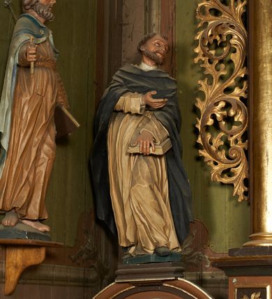 Zdjęcie nr 2: Figura drążona z tyłu, ustawiona na niewielkim postumencie. Święty ukazany frontalnie, pochylony w lewo, z uniesioną głową, w silnym kontrapoście. Lewą ręką przytrzymuje z przodu, na udzie księgę, prawą ugiętą, kieruje w lewo. Twarz ma szczupłą, o głęboko osadzonych, dużych oczach, skierowanych w górę, dużym nosie oraz wąskich, rozchylonych ustach. Twarz okolona jest krótką brodą oraz krótkimi włosami, na głowie tonsura. Święty ubrany w obficie fałdowany, biały habit dominikański oraz czarny płaszcz z kapturem. Polichromia ciała naturalistyczna.