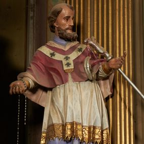 Zdjęcie nr 1: Figura drążona z tyłu, ustawiona na niewielkim prostopadłościennym postumencie. Święty ukazany jest frontalnie, lekko odchylony, z głową skierowaną w lewo, w kontrapoście. Obie ręce ma rozłożone na boki, w prawej dłoni trzyma kamień (?). Twarz ma szczupłą, wyciągniętą, o wysokim czole, silnie zaznaczonych kościach policzkowych; ma duże oczy, lekko garbaty nos oraz wąskie, rozchylone usta. Twarz okolona jest gęstą, brązową brodą, na głowie ma łysinę, po bokach głowy i nad czołem kręcone włosy. Święty ubrany jest w jasnofioletową sutannę, białą, złoconą przy brzegu komżę, czerwoną pelerynkę z różową podszewką oraz paliusz. Polichromia naturalistyczna w partiach ciała.