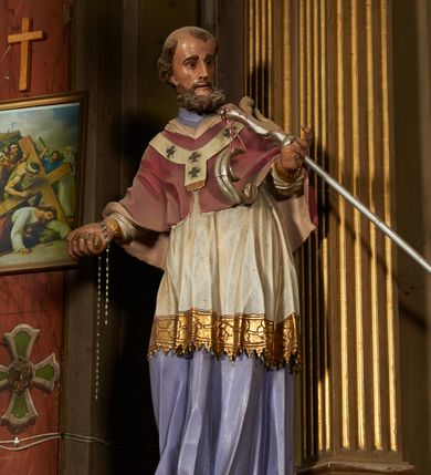 Zdjęcie nr 3: Figura drążona z tyłu, ustawiona na niewielkim prostopadłościennym postumencie. Święty ukazany jest frontalnie, lekko odchylony, z głową skierowaną w lewo, w kontrapoście. Obie ręce ma rozłożone na boki, w prawej dłoni trzyma kamień (?). Twarz ma szczupłą, wyciągniętą, o wysokim czole, silnie zaznaczonych kościach policzkowych; ma duże oczy, lekko garbaty nos oraz wąskie, rozchylone usta. Twarz okolona jest gęstą, brązową brodą, na głowie ma łysinę, po bokach głowy i nad czołem kręcone włosy. Święty ubrany jest w jasnofioletową sutannę, białą, złoconą przy brzegu komżę, czerwoną pelerynkę z różową podszewką oraz paliusz. Polichromia naturalistyczna w partiach ciała.