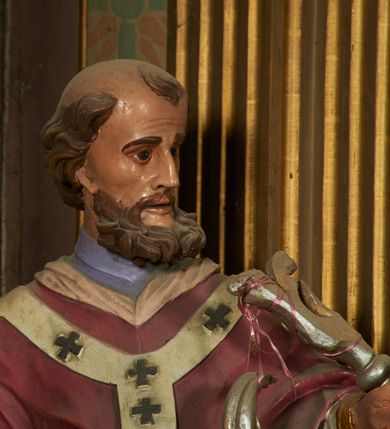 Zdjęcie nr 4: Figura drążona z tyłu, ustawiona na niewielkim prostopadłościennym postumencie. Święty ukazany jest frontalnie, lekko odchylony, z głową skierowaną w lewo, w kontrapoście. Obie ręce ma rozłożone na boki, w prawej dłoni trzyma kamień (?). Twarz ma szczupłą, wyciągniętą, o wysokim czole, silnie zaznaczonych kościach policzkowych; ma duże oczy, lekko garbaty nos oraz wąskie, rozchylone usta. Twarz okolona jest gęstą, brązową brodą, na głowie ma łysinę, po bokach głowy i nad czołem kręcone włosy. Święty ubrany jest w jasnofioletową sutannę, białą, złoconą przy brzegu komżę, czerwoną pelerynkę z różową podszewką oraz paliusz. Polichromia naturalistyczna w partiach ciała.