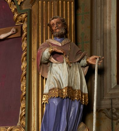 Zdjęcie nr 2: Figura drążona od tyłu, ustawiona na niewielkim prostopadłościennym postumencie. Święty ukazany jest frontalnie, w kontrapoście, o odchylonej sylwetce, z głową skierowaną w prawo. W lewej, ugiętej i odchylonej ręce trzyma laskę, prawą kładzie na piersi. Twarz ma szczupłą, podłużną o silnie zaznaczonych kościach policzkowych, dużych, migdałowatych oczach, lekko garbatym nosie i długich, wąskich, rozchylonych ustach, okoloną gęstą, brązową, kręconą brodą i kręconymi włosami. Święty ubrany jest w jasnofioletową sutannę, białą, złoconą przy brzegu komżę, czerwoną pelerynkę o różowej podszewce. Na piersi ma zawieszony pektorał. Przy lewej nodze biała, lamowana infuła ze złoconym krzyżem. Polichromia ciała naturalistyczna. 
