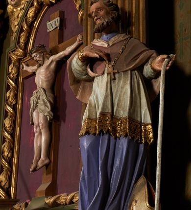 Zdjęcie nr 4: Figura drążona od tyłu, ustawiona na niewielkim prostopadłościennym postumencie. Święty ukazany jest frontalnie, w kontrapoście, o odchylonej sylwetce, z głową skierowaną w prawo. W lewej, ugiętej i odchylonej ręce trzyma laskę, prawą kładzie na piersi. Twarz ma szczupłą, podłużną o silnie zaznaczonych kościach policzkowych, dużych, migdałowatych oczach, lekko garbatym nosie i długich, wąskich, rozchylonych ustach, okoloną gęstą, brązową, kręconą brodą i kręconymi włosami. Święty ubrany jest w jasnofioletową sutannę, białą, złoconą przy brzegu komżę, czerwoną pelerynkę o różowej podszewce. Na piersi ma zawieszony pektorał. Przy lewej nodze biała, lamowana infuła ze złoconym krzyżem. Polichromia ciała naturalistyczna. 
