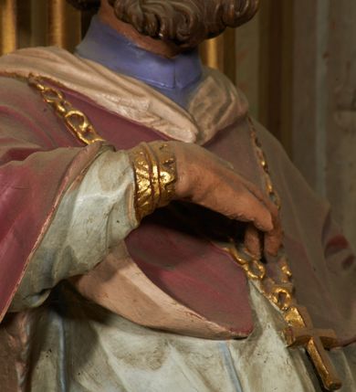 Zdjęcie nr 8: Figura drążona od tyłu, ustawiona na niewielkim prostopadłościennym postumencie. Święty ukazany jest frontalnie, w kontrapoście, o odchylonej sylwetce, z głową skierowaną w prawo. W lewej, ugiętej i odchylonej ręce trzyma laskę, prawą kładzie na piersi. Twarz ma szczupłą, podłużną o silnie zaznaczonych kościach policzkowych, dużych, migdałowatych oczach, lekko garbatym nosie i długich, wąskich, rozchylonych ustach, okoloną gęstą, brązową, kręconą brodą i kręconymi włosami. Święty ubrany jest w jasnofioletową sutannę, białą, złoconą przy brzegu komżę, czerwoną pelerynkę o różowej podszewce. Na piersi ma zawieszony pektorał. Przy lewej nodze biała, lamowana infuła ze złoconym krzyżem. Polichromia ciała naturalistyczna. 

