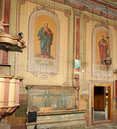 Zdjęcie nr 2: Polichromia ścian kościoła odpowiada podziałom architektonicznym wnętrza wydzielonym przez pilastry, filary i kolumny (w prezbiterium iluzjonistyczne pilastry). Malowany jasnobrązowy cokół w jednej czwartej wysokości ścian zamknięty jest profilowanym gzymsem, w jego polu znajdują się jasnozielone malowane płyciny odpowiadające szerokości podziałów wnętrza. Trzony pilastrów bladoróżowe z malowanymi czerwonymi płycinami, w prezbiterium z kapitelami korynckimi, w nawie zakończone tryglifami. Podpory podtrzymują malowane belkowanie, z jasnoróżowym fryzem i wspornikami dekorowanymi anthemionami, dźwigającymi silnie wysunięty gzyms. W nawie belkowanie malowane jest w partii środkowej (bez zaskrzynień) podtrzymywanej przez filary i kolumny. W polach pomiędzy pilastrami malowane płyciny w formie prostokątów, o szerokości przęseł, ujęte ramą dekorowaną w narożach stylizowanym akantem z kampanulami. W prezbiterium w jasnożółtych płycinach znajdują się malowane nisze w formie stojącego prostokąta zamkniętego łukiem półkolistym, ujęte profilowaną ramą, dekorowaną u góry i u dołu ornamentem roślinnym, z przedstawieniami ewangelistów i ich symboli. Na ścianie północnej znajdują się św. Mateusz, św. Marek i św. Łukasz, a na ścianie południowej, pomiędzy okami św. Jan. W nawie jasnożółte płyciny ścian wypełnione są dekoracją geometryczną w formie niebieskich i żółtych równoramiennych krzyżyków. Otwory okienne, drzwiowe i arkada kaplicy ujęte są malowanymi, imitującymi profilowanie obramieniami, dekorowanymi ornamentem roślinnym. 
Strop prezbiterium i nawy w malowanej, kolorowej, profilowanej ramie, po wewnętrznej stronie z zielono-różowym pasem zdobionym ornamentem. Pośrodku, na tle malowanej jasnoróżowej płyciny, wydzielone pole o fantazyjnym kształcie, obwiedzione pomarańczową opaską z kartuszami na każdej stronie, ujętymi ornamentem roślinnym, wypełnionymi kratką i fantazyjną dekoracją. Jasnoniebieskie pole wypełnione jest malowanymi na złoto gwiazdami. W prezbiterium, w jego centrum kolejne pole o analogicznym kształcie ze sceną Wniebowzięcia Marii, a w nawie dwa pola ze scenami Chrystusa Zmartwychwstałego i Świętej Rodziny. W partii zaskrzynień nawy malowane płyciny w zielonych opaskach i jasnoróżowych polach wypełnione ornamentem geometrycznym. Jasnozielona balustrada chóru muzycznego dekorowana malowanymi płycinami w formie prostokątów, naprzemiennie stojących i leżących, flankujących środkowy medalion, wszystkie ujęte profilowaną ramą. Pola prostokątów stojących wypełnione rozetami na jasnoczerwonym tle. Pola prostokątów leżących wypełnione przedstawieniami instrumentów i ornamentu roślinnego ukazanych na żółtym tle. W środkowym medalionie przedstawienie Dawida grającego na harfie. 















