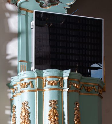 Zdjęcie nr 2: Ambona przyścienna, wisząca z dostępem od strony prezbiterium. Kosz na planie ośmioliścia z trójkątnymi wypustkami pomiędzy ściankami oraz dodatkowym podestem od strony prezbiterium na planie połowy z sześcioboku, do którego prowadzą schody z pełną balustradą. Spód kosza puklowany, zakończony szyszką. Wypukłe ścianki kosza dekorowane płaskorzeźbionymi, owocowymi girlandami podwieszonymi na wstędze; w górnej partii pas wydzielony dwiema listewkami z kwiatowymi girlandami, opasujący kosz i podest. Parapet w formie profilowanego gzymsu. Zaplecek wąski, ujęty pilastrami na kanelowanych cokołach. Baldachim na planie czworoliścia z trójkątnymi wypustkami pomiędzy ściankami; w formie niepełnego belkowania; fryz dekorowany kwiatowymi girlandami. Na baldachimie dwa akroteriony. U spodu czworolistna płycina z rzeźbioną gołębicą Ducha Świętego. Struktura polichromowana w odcieniu bladozielonym, ornamentyka i profile złocone, gołębica srebrzona.