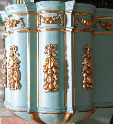 Zdjęcie nr 1: Ambona przyścienna, wisząca z dostępem od strony prezbiterium. Kosz na planie ośmioliścia z trójkątnymi wypustkami pomiędzy ściankami oraz dodatkowym podestem od strony prezbiterium na planie połowy z sześcioboku, do którego prowadzą schody z pełną balustradą. Spód kosza puklowany, zakończony szyszką. Wypukłe ścianki kosza dekorowane płaskorzeźbionymi, owocowymi girlandami podwieszonymi na wstędze; w górnej partii pas wydzielony dwiema listewkami z kwiatowymi girlandami, opasujący kosz i podest. Parapet w formie profilowanego gzymsu. Zaplecek wąski, ujęty pilastrami na kanelowanych cokołach. Baldachim na planie czworoliścia z trójkątnymi wypustkami pomiędzy ściankami; w formie niepełnego belkowania; fryz dekorowany kwiatowymi girlandami. Na baldachimie dwa akroteriony. U spodu czworolistna płycina z rzeźbioną gołębicą Ducha Świętego. Struktura polichromowana w odcieniu bladozielonym, ornamentyka i profile złocone, gołębica srebrzona.