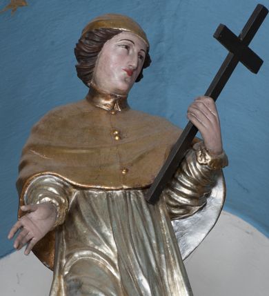 Zdjęcie nr 1: Rzeźba przedstawiająca św. Karola Boromeusza, ścięta z tyłu, ustawiona na niskim cokole. Figura zwrócona delikatnie w prawo, z prawą nogą ugiętą w kolanie, z głową skierowaną w lewo. Obie dłonie wyciąga przed siebie, w lewej trzyma krzyż. Twarz pełna, z długim nosem, wąskimi ustami i małymi oczami, włosy krótkie, zasłaniające uszy, zawinięte do tyłu w bujne loki. Święty ubrany jest w złoconą albę, srebrzoną rokietę oraz złocony mucet, na głowie ma założoną złoconą piuskę, na stopach złocone buty. Polichromia naturalistyczna w odsłoniętych partiach ciała, szaty złocone i srebrzone.
