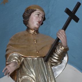 Zdjęcie nr 1: Rzeźba przedstawiająca św. Karola Boromeusza, ścięta z tyłu, ustawiona na niskim cokole. Figura zwrócona delikatnie w prawo, z prawą nogą ugiętą w kolanie, z głową skierowaną w lewo. Obie dłonie wyciąga przed siebie, w lewej trzyma krzyż. Twarz pełna, z długim nosem, wąskimi ustami i małymi oczami, włosy krótkie, zasłaniające uszy, zawinięte do tyłu w bujne loki. Święty ubrany jest w złoconą albę, srebrzoną rokietę oraz złocony mucet, na głowie ma założoną złoconą piuskę, na stopach złocone buty. Polichromia naturalistyczna w odsłoniętych partiach ciała, szaty złocone i srebrzone.
