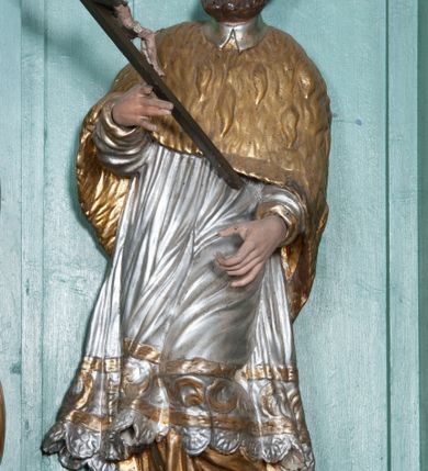 Zdjęcie nr 2: Rzeźba przedstawiająca św. Jana Nepomucena, ścięta z tyłu, ustawiona na rzeźbionym fragmencie skały. Figura zwrócona trzy czwarte w prawo, z lewą nogą ugiętą w kolanie i wspartą na skale, z głową skierowaną w prawo. Obie dłonie wyciąga przed siebie: w prawej trzyma krucyfiks, lewą ma ułożoną do trzymania niezachowanego atrybutu (zapewne palmy męczeństwa). Twarz szeroka, okolona krótką brodą, włosy krótkie, zasłaniające uszy, zawinięte do tyłu w bujne loki. Ubrany jest w złoconą albę, srebrzoną rokietę oraz złoconą almucję, na głowie ma założony czarny biret, na stopach złocone buty. Polichromia naturalistyczna w odsłoniętych partiach ciała, szaty złocone i srebrzone.