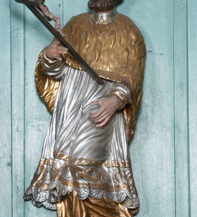 Zdjęcie nr 3: Rzeźba przedstawiająca św. Jana Nepomucena, ścięta z tyłu, ustawiona na rzeźbionym fragmencie skały. Figura zwrócona trzy czwarte w prawo, z lewą nogą ugiętą w kolanie i wspartą na skale, z głową skierowaną w prawo. Obie dłonie wyciąga przed siebie: w prawej trzyma krucyfiks, lewą ma ułożoną do trzymania niezachowanego atrybutu (zapewne palmy męczeństwa). Twarz szeroka, okolona krótką brodą, włosy krótkie, zasłaniające uszy, zawinięte do tyłu w bujne loki. Ubrany jest w złoconą albę, srebrzoną rokietę oraz złoconą almucję, na głowie ma założony czarny biret, na stopach złocone buty. Polichromia naturalistyczna w odsłoniętych partiach ciała, szaty złocone i srebrzone.