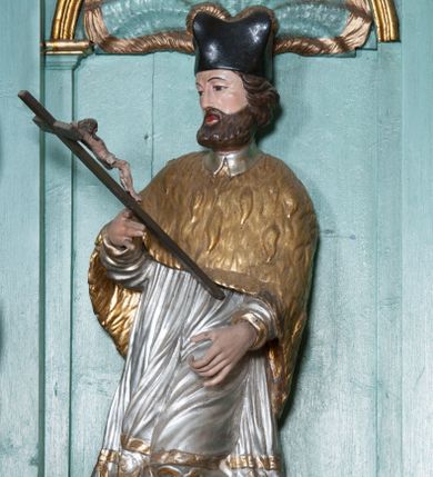 Zdjęcie nr 1: Rzeźba przedstawiająca św. Jana Nepomucena, ścięta z tyłu, ustawiona na rzeźbionym fragmencie skały. Figura zwrócona trzy czwarte w prawo, z lewą nogą ugiętą w kolanie i wspartą na skale, z głową skierowaną w prawo. Obie dłonie wyciąga przed siebie: w prawej trzyma krucyfiks, lewą ma ułożoną do trzymania niezachowanego atrybutu (zapewne palmy męczeństwa). Twarz szeroka, okolona krótką brodą, włosy krótkie, zasłaniające uszy, zawinięte do tyłu w bujne loki. Ubrany jest w złoconą albę, srebrzoną rokietę oraz złoconą almucję, na głowie ma założony czarny biret, na stopach złocone buty. Polichromia naturalistyczna w odsłoniętych partiach ciała, szaty złocone i srebrzone.