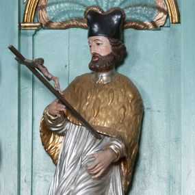 Zdjęcie nr 1: Rzeźba przedstawiająca św. Jana Nepomucena, ścięta z tyłu, ustawiona na rzeźbionym fragmencie skały. Figura zwrócona trzy czwarte w prawo, z lewą nogą ugiętą w kolanie i wspartą na skale, z głową skierowaną w prawo. Obie dłonie wyciąga przed siebie: w prawej trzyma krucyfiks, lewą ma ułożoną do trzymania niezachowanego atrybutu (zapewne palmy męczeństwa). Twarz szeroka, okolona krótką brodą, włosy krótkie, zasłaniające uszy, zawinięte do tyłu w bujne loki. Ubrany jest w złoconą albę, srebrzoną rokietę oraz złoconą almucję, na głowie ma założony czarny biret, na stopach złocone buty. Polichromia naturalistyczna w odsłoniętych partiach ciała, szaty złocone i srebrzone.