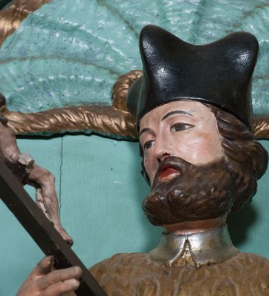 Zdjęcie nr 4: Rzeźba przedstawiająca św. Jana Nepomucena, ścięta z tyłu, ustawiona na rzeźbionym fragmencie skały. Figura zwrócona trzy czwarte w prawo, z lewą nogą ugiętą w kolanie i wspartą na skale, z głową skierowaną w prawo. Obie dłonie wyciąga przed siebie: w prawej trzyma krucyfiks, lewą ma ułożoną do trzymania niezachowanego atrybutu (zapewne palmy męczeństwa). Twarz szeroka, okolona krótką brodą, włosy krótkie, zasłaniające uszy, zawinięte do tyłu w bujne loki. Ubrany jest w złoconą albę, srebrzoną rokietę oraz złoconą almucję, na głowie ma założony czarny biret, na stopach złocone buty. Polichromia naturalistyczna w odsłoniętych partiach ciała, szaty złocone i srebrzone.