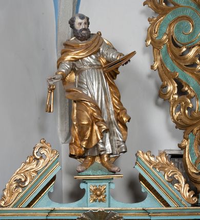Zdjęcie nr 2: Rzeźba przedstawiająca św. Piotra, ścięta z tyłu, drążona, ustawiona na rzeźbionym fragmencie skały. Figura ukazana frontalnie z głową skierowaną delikatnie w prawo, obie ręce rozłożone w boki: w lewej trzyma zamkniętą księgę, a w prawej klucze. Twarz szeroka z otwartymi ustami, okolona gęstą brodą, włosy w postaci pukli po bokach i na czubku głowy. Święty ubrany jest w delikatnie drapowaną suknię, przepasaną w talii oraz płaszcz zapięty na lewym ramieniu, opadający na plecy i na prawe kolano, drapowany w grube fałdy, zawijające się przy brzegach; na stopach ma sandały. Polichromia naturalistyczna w odsłoniętych partiach ciała, szaty i atrybuty złocone oraz srebrzone.